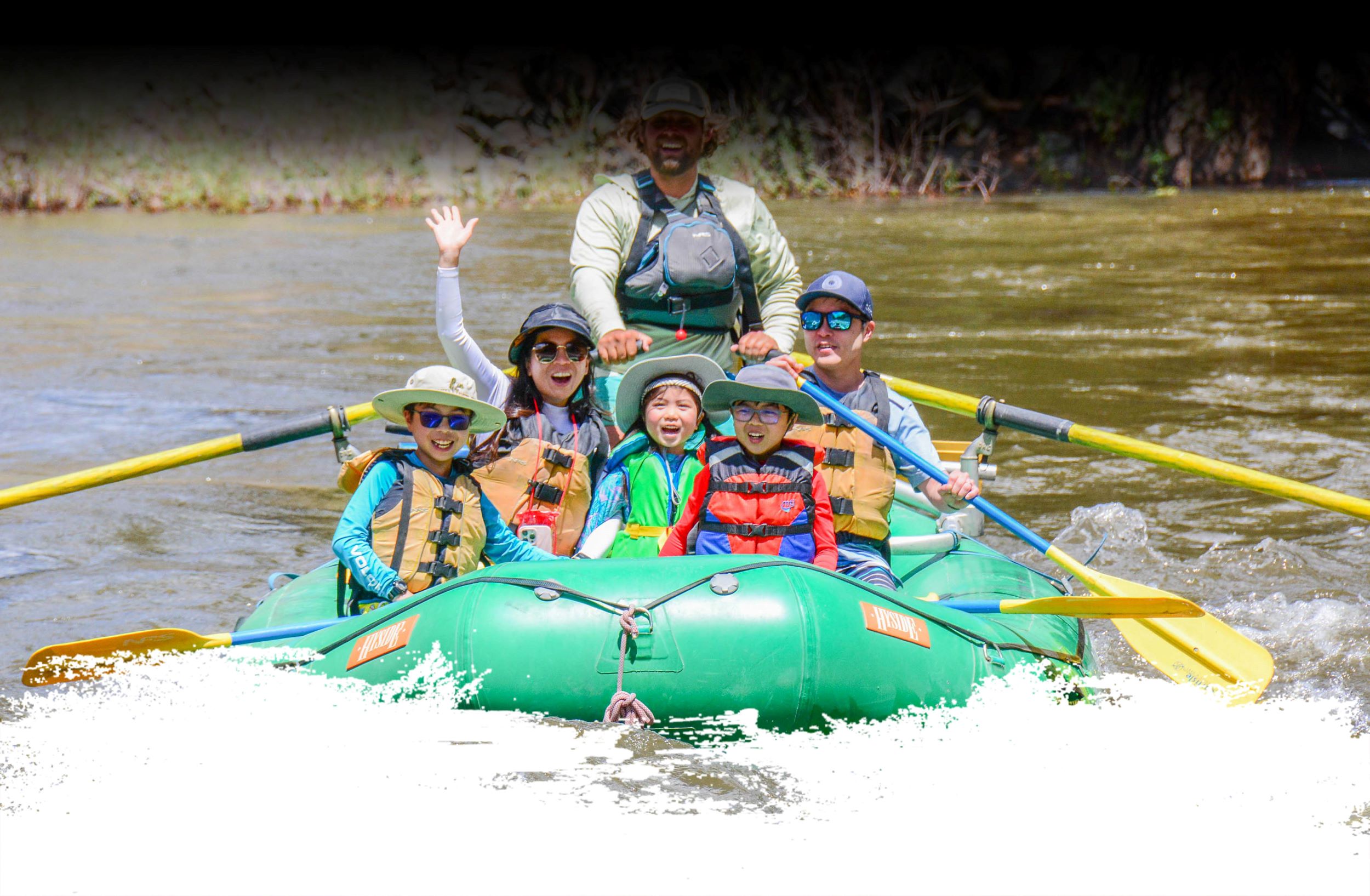 A smiling family paddles together down a calm stretch of river in a bright raft, surrounded by sunshine and mountain views—capturing the joy of a family friendly white water rafting trip in Colorado.